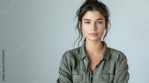 A portrait of a young woman with natural makeup, wearing a casual olive green shirt, looking directly at the camera with a neutral expression against a plain grey background.