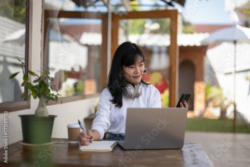 Young woman wearing headphones holds a notebook and uses a laptop at an outdoor cafe table. Finding and thinking of ideas, writing them down in a notebook