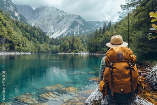 A hiker with a backpack sits lakeside, staring at a clear mountain lake and lush greenery of a forest