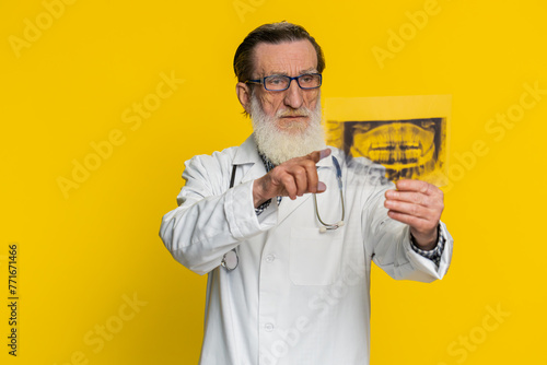 Senior elderly doctor orthodontist man examines a panoramic x-ray picture of the jaw teeth. 3D model of the patient's mouth, MRI scan. Dentistry oral care. Stomatology grandfather on yellow background