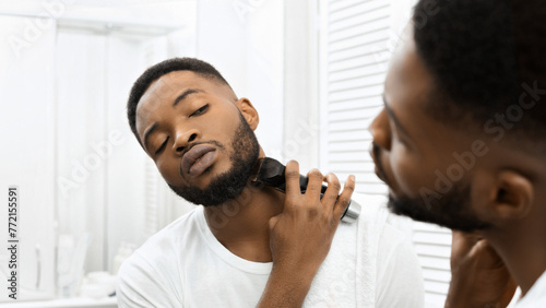 Man trimming beard with electric clippers in mirror in bathroom
