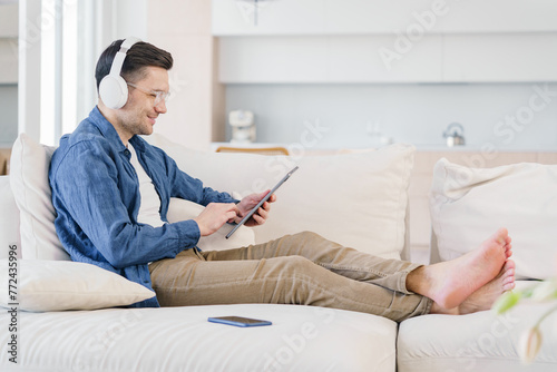 A relaxed man in headphones enjoys using a tablet while reclining on a plush sofa, immersed in a digital world within his bright living space.