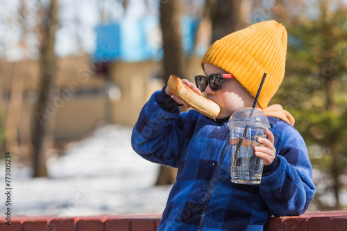 A little boy eats fast food outside in the spring. He will rub the baby in a fashionable plaid shirt, yellow hat and sunglasses. The child eats a hot dog and drinks lemonade.