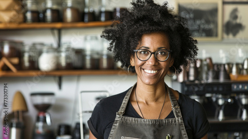 A woman with curly hair and glasses is smiling in front of a counter with jars