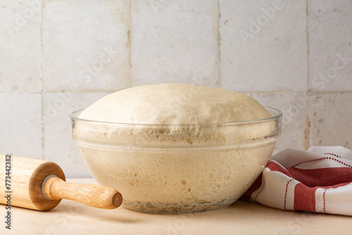 Glass bowl with fresh homemade yeast dough on a kitchen table with utensils.