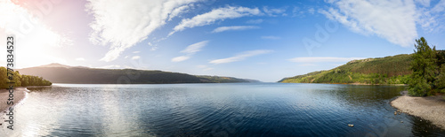 Panoramic drone shot of Loch Ness, where the gentle rays of the sun bathe the tranquil waters and pebbled shores in a warm glow, with dense forests and hills framing the scene in natural splendor