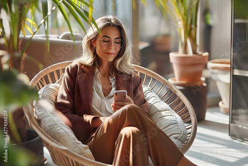 Beschäftigte, schöne, reife Geschäftsfrau mittleren Alters, die ihr Mobiltelefon benutzt und auf ihr Smartphone schaut, sitzt in einem bequemen Stuhl in einem modernen Büro mit grünen Pflanzen
