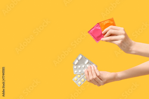 Female hands with birth control pills and condoms on yellow background