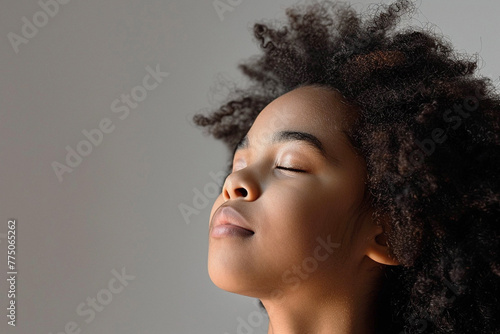 Photo of a beautiful African American teenage girl with black short wavy hair inhaling air with her eyes closed against a light background.
