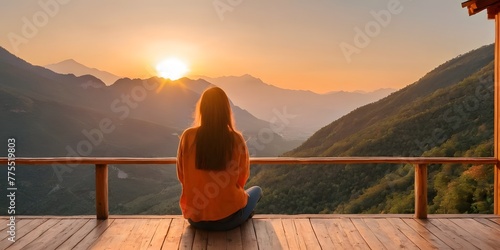 Back view of a woman sitting on wooden porch extending into a high mountain cliff. The sun is setting on the mountain and there is a beautiful warm view