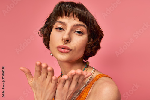 flirty young woman with nose piercing and short brunette hair sending air kiss on pink background