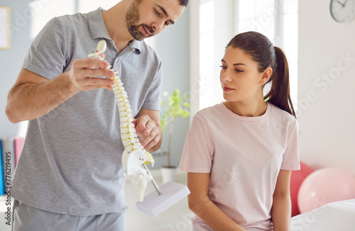 Portrait of male doctor physiotherapist or orthopedist showing to his woman patient model of spine in rehab clinic giving consultation about scoliosis or spinal problems during medical exam.