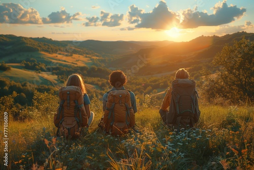 Three individuals with backpacks are perched atop a hill, gazing at the sunset over the natural landscape. The sky is painted with vibrant colors, casting a warm glow on the grassland below