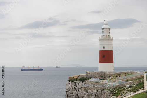 A lighthouse is on a rocky cliff overlooking the ocean. The lighthouse is red and white. A large ship is in the distance