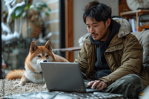 A man is comfortably sitting on the floor next to a window, using a personal computer, with a carnivore companion dog laying next to him