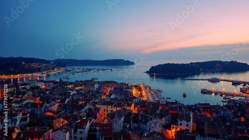Birds eye view of the evening city during sunset. Aerial photography. Lights on lanterns on dusk. The ships are in the bay. Tiled roofs of houses in the old town. Rovinj, Croatia - April 7, 2024