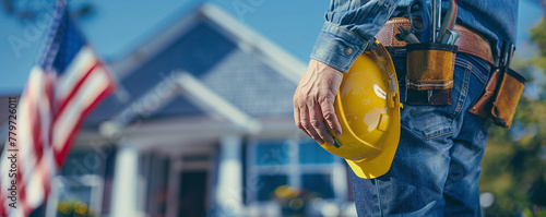 hand of an American worker in blue jeans holding a yellow helmet on a tool belt on which tools hang at shoulder level, blurred background of a house