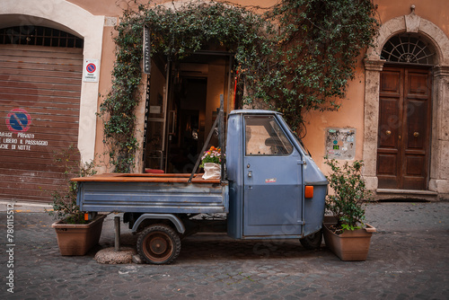 Charming urban scene in Italy with an Ape truck parked on cobblestone street. Detailed architectural elements, shops, and signage add picturesque charm.