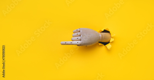 A wooden hand protrudes from a torn hole in yellow paper and points its index finger to the left. Concept of direction, adjustment and assistance. Mannequin, robot