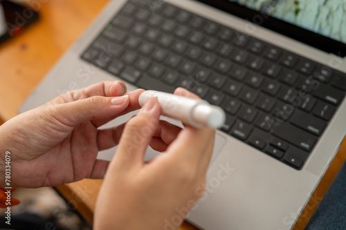 Close-up of hands using a portable lancet device for blood sugar testing over a laptop.
