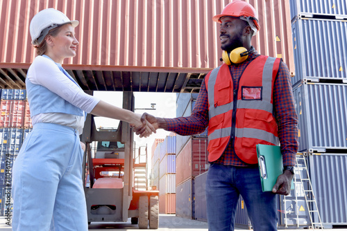 Two worker wearing safety helmet shaking hands at logistic shipping cargo containers yard. African American engineer man having hand shack with his beautiful young woman boss after work discussion.