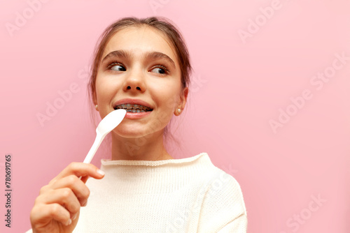 hungry teenage girl with braces licks spoon and dreams on pink isolated background, pensive child bites spoon with teeth and plans and imagines