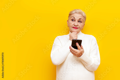thoughtful old grandmother in a white sweater uses a smartphone and thinks and plans on a yellow isolated background, elderly woman pensioner holds a mobile and imagines
