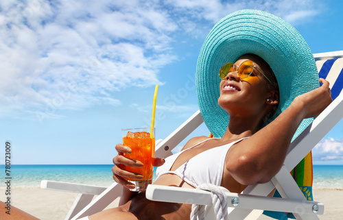 Happy woman is sunbathing on a beach deck chair, wearing sun hat and sunglasses, drinking a orange juice on a sunny day by the seaside, concept of a summer beach holiday, booking travel and resort