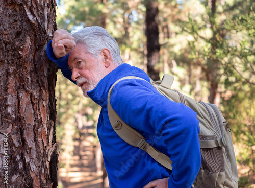 An active senior man with backpack breathless on a day hike in a mountain forest leans against a tree trunk to regain his strength