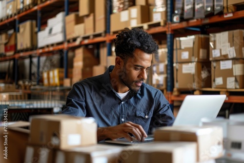 A young man in a warehouse concentrating on work at his laptop, surrounded by boxes and shelves of inventory