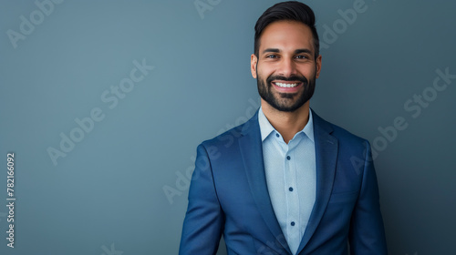 A confident Indian businessman in his late 40s, wearing a blue suit, standing against a grey background and smiling for the camera.