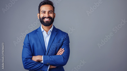 A confident Indian businessman in his late 40s, wearing a blue suit, standing against a grey background and smiling for the camera.