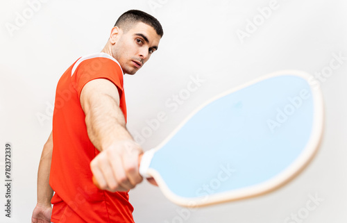 A shaved man in his 30s poses on a white background with a pickleball racket in sports clothing. The young man is backhanding the racket towards the camera. Pickle ball concept.