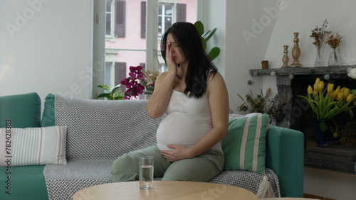 Pregnant woman having ache, 30s lady struggling with nausea during third trimester of pregnancy seated on couch sofa at home residence gently touching belly