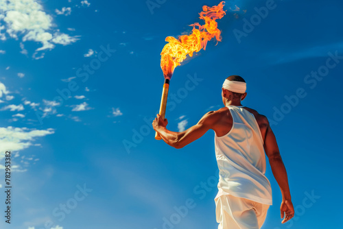 An athlete in white clothes with an olympic torch stands solemnly against the blue sky. Back view. space for copying.