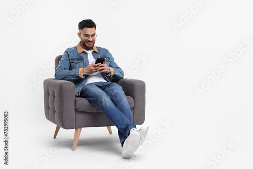 Happy man using smartphone in armchair on white background, space for text