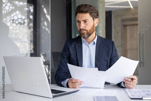 A serious young man businessman works in the office at a desk, looks at the screen of a laptop, holds documents in his hands, compares data, makes statistics and analysis