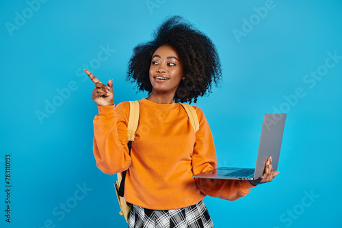 An African American college girl in casual attire holding a laptop, pointing to the side in a studio with a blue backdrop.