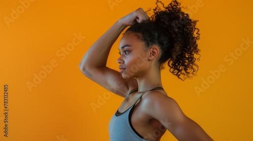 Focused young woman with curly hair displaying strength, flexing biceps in front of a yellow wall