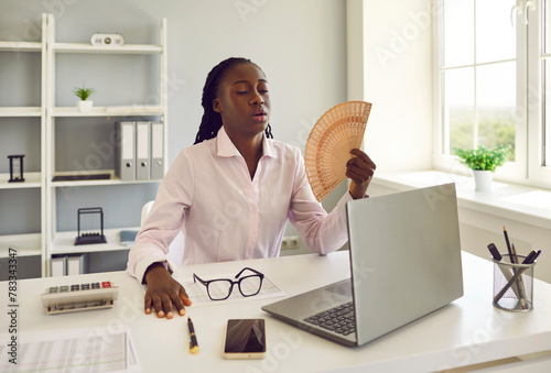 Young african american business woman employee using hand fan suffering from heat and summer high temperature working at the desk in office at her workplace. Heatwave problem in office.