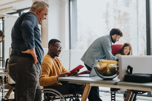 An inclusive modern office environment with a diverse team collaborating. A person in a wheelchair actively engages with colleagues.