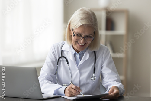 Mature woman doctor sit at desk make notes, jotting information, prepare paper document, reviewing medical records, completing administrative tasks, filling forms or writing prescriptions at workplace
