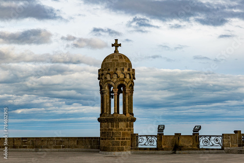 Stone bell tower in neo Gothic style of Sagrat Cor church on top of Mount Tibidabo, cross on small dome, upper esplanade, cloudy day with blue sky with clouds in Barcelona, Catalonia, Spain