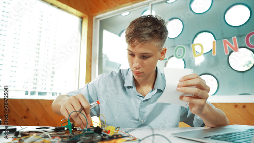 Smart boy inspect or learning to use electronic tool while looking at note. Highschool student studying or inspecting about electric equipment while sitting at STEM technology class. Edification.