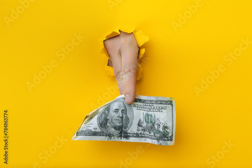 A right man's hand holds dirty money through a torn hole in yellow paper. Concept of dishonest income, donation, profit and salary fraud.