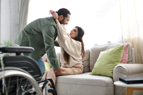 loving man in homewear helping his wife with mobility disability to get to couch from wheelchair