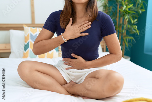 Young asian woman sitting on bed with hands on chest and stomach at bedroom