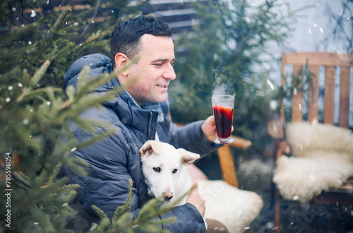 Happy man sitting on wooden chair with dog on laps in backyard, drinking hot aromatic grog in winter