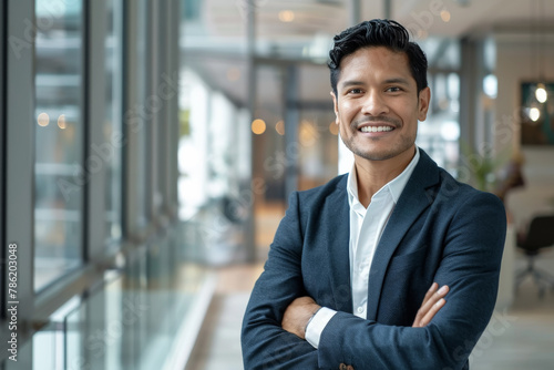 Atractivo hombre joven latino americano de negocios con traje azul marino, brazos cruzados, con fondo desenfocado de instalaciones empresariales modernas y luminosas