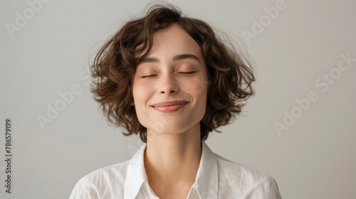 Portrait of a shy female corporate intern smiling with eyes shut against grey background